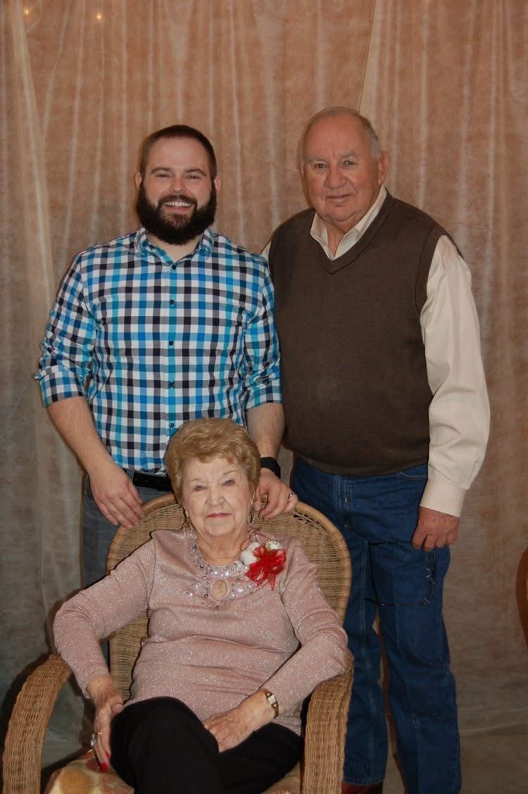 Blake and Daddy Bob stand behind Mee Maw, who sits in a wicker chair in a pink top with a red corsage, in front of a draped curtain.