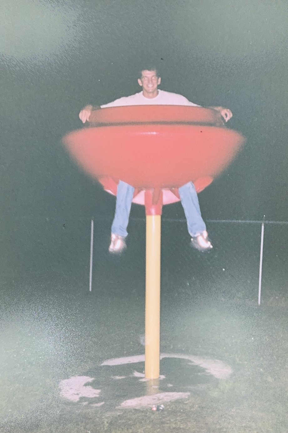 A night photograph taken with a flash: a smiling young man in a white T-shirt and jeans sits in a red bowl-shaped seat on top of a yellow pole, his arms over the rim and his legs hanging below it. Behind him, a fence stands in the dark.